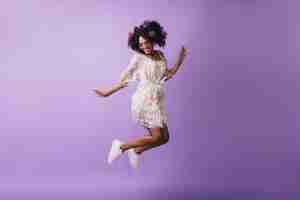 Free photo carefree african girl in white shoes jumping. adorable female model with flowers in hair dancing with happy smile.