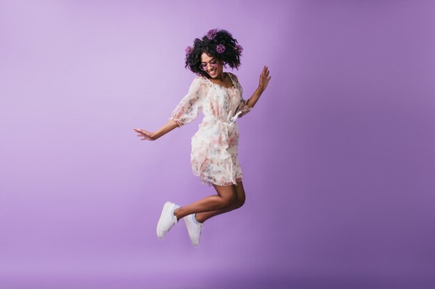 흰색 신발 점프에 평온한 아프리카 소녀입니다. 행복 한 미소와 함께 춤을 머리에 꽃을 가진 사랑스러운 여성 모델.