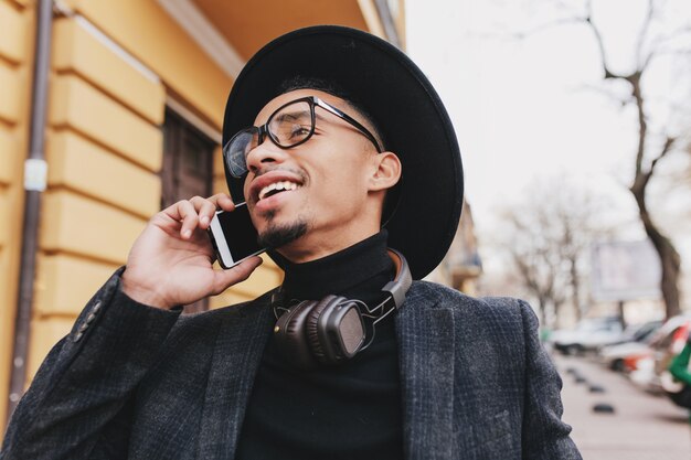 Carefree african guy with short haircut talking on phone with smile. Outdoor photo of enthusiastic black young man in hat walking down the street with cell.