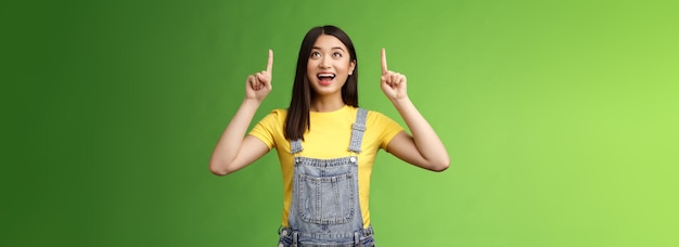 Carefree amused asian brunette in yellow tshirt dungarees smiling amazed and fascinated look pointin