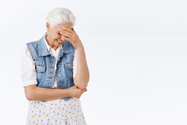 Free photo carefree amused charming modern granny with grey combed hair in stylish denim vest dress laughing over hilarious kid outfit or joke look down chuckling and smiling face palm touch forehead