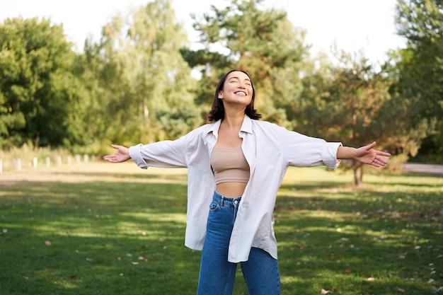 Carefree asian girl laughing and dancing in park enjoying summer sunny day raising hands up and brea