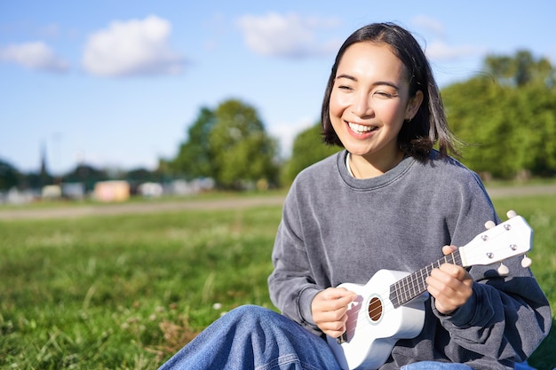 Free photo carefree asian girl singing and playing ukulele in park sitting on grass musician relaxing on her free time outdoors