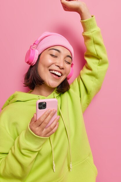 Carefree Asian woman has upbeat mood dances with rhythm of music uses modern cellular and headphones wears hat and sweatshirt isolated over pink wall spends leisure time with favorite playlist