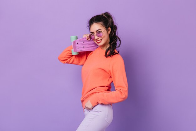 Carefree asian woman with ponytail holding skateboard. Studio shot of smiling young woman with longboard isolated on purple background.