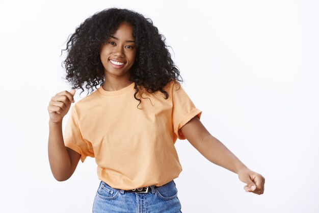 평온하고 매력적인 젊은 아프리카계 미국인 곱슬머리 소녀가 봄 주황색 티셔츠를 입고 손을 흔들며 춤을 추며 활짝 웃는 멋진 파티를 즐기고 즐거운 흰색 벽을 즐기고 있습니다.
