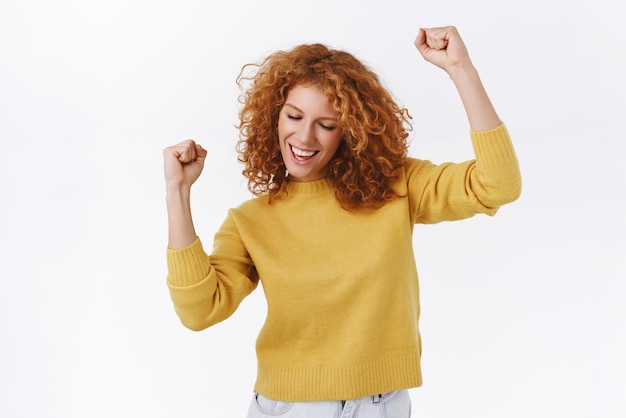 Carefree attractive modern redhead girl in yellow sweater dancing as triumphing feeling joy of success delighted moving hands fist pump close eyes and smiling pleased white background