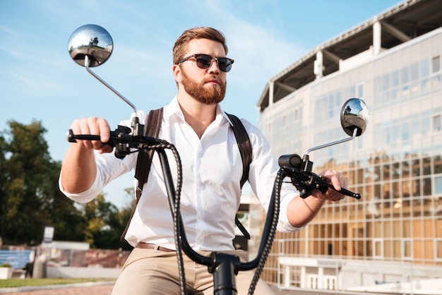 Беззаботный бородатый мужчина в солнцезащитных очках с рюкзаком едет на современном мотоцикле на улице и смотрит в сторону