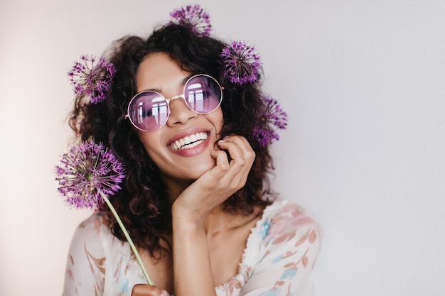 Bella ragazza africana spensierata in posa con fiori e sorridente foto interna di un'incredibile modella femminile bruna che esprime emozioni positive