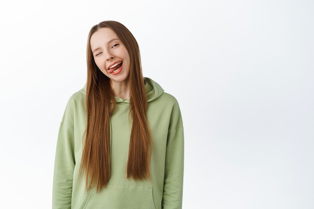 Carefree beautiful millennial girl show tongue, smiling with white perfect teeth and winking, standing in hoodie with positive emotion, studio wall