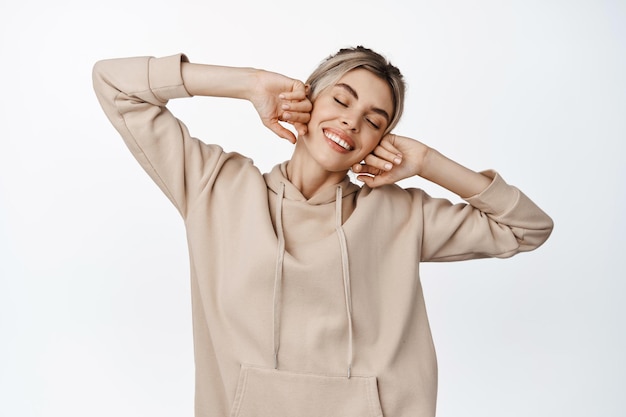 Carefree beautiful woman holding hands near natural clean face close eyes and smiling happy standing in beige hoodie over white background