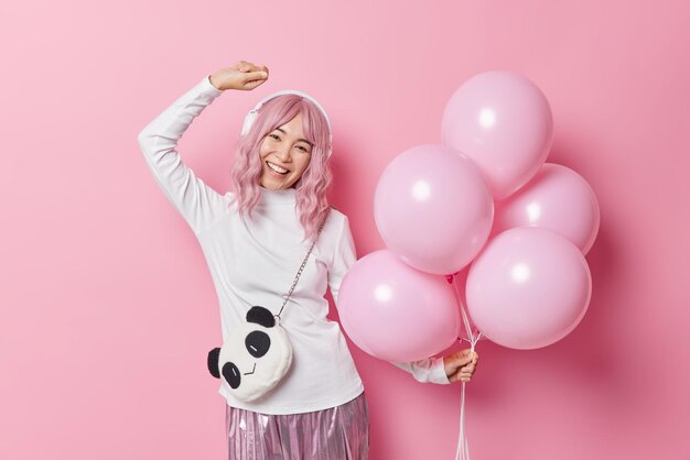Carefree birthday girl dances with rhythm of music listens audio track via headphones holds bunch of inflated balloons isolated over pink background enjoys party time. People and celebration