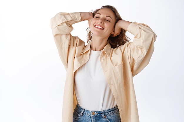 평온한 금발 여성은 편안하고, 머리를 만지고, 자유롭고 행복하며, 눈을 감고, 여가를 즐기고, 흰 벽에 기대어 서 있습니다.