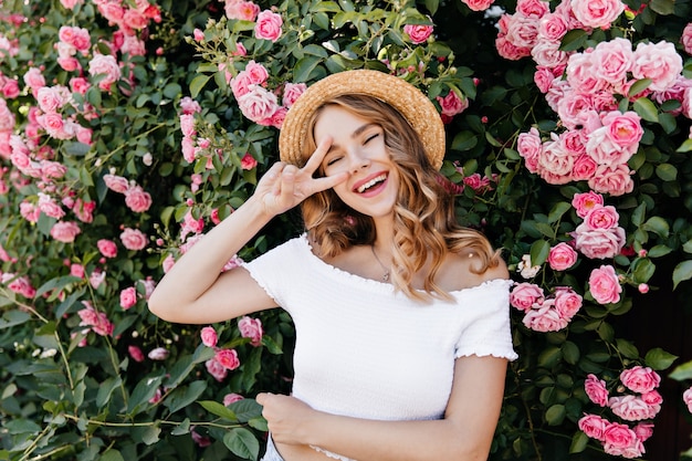 Carefree blonde girl laughing while posing in garden. Outdoor portrait of adorable woman in hat surrounded by roses.