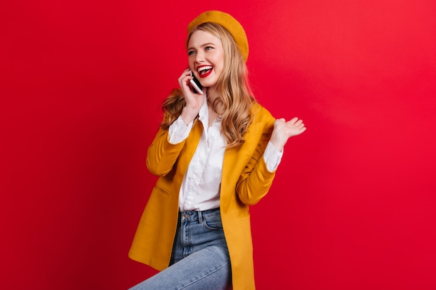 Carefree blonde girl talking on phone and dancing.  fashionable french woman in beret holding smartphone on red wall.