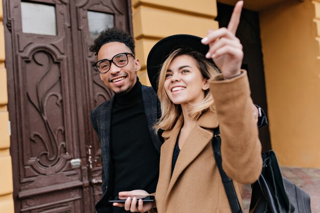 Carefree blonde woman in hat showing her african friend something interesting. Outdoor portrait of smiling black guy in glasses walking around town in cold day with fair-haired lady.