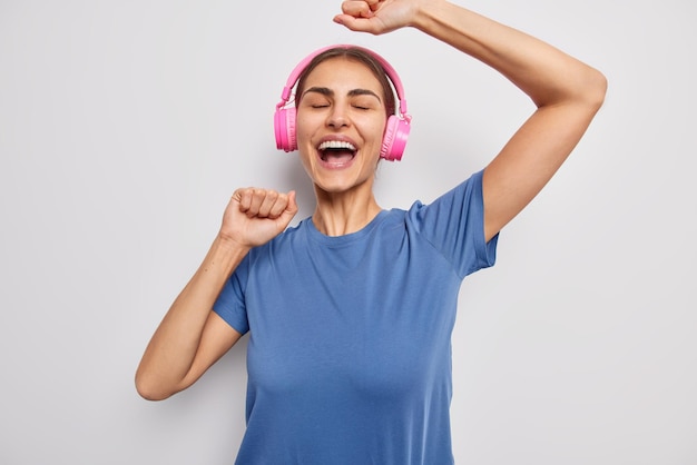 Free photo carefree brunette woman dances with rhythm of music wears stereo headphones on ears shakes arms smiles broadly dressed in casual blue t shirt isolated over white background positive meloman