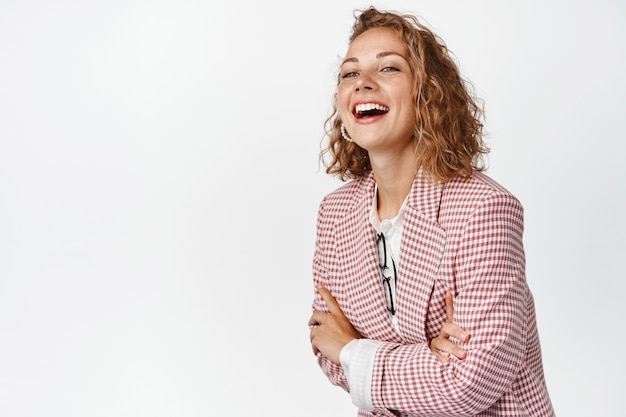 Carefree business woman laughing, cross arms on chest, smiling and watching something funny, standing in suit on white