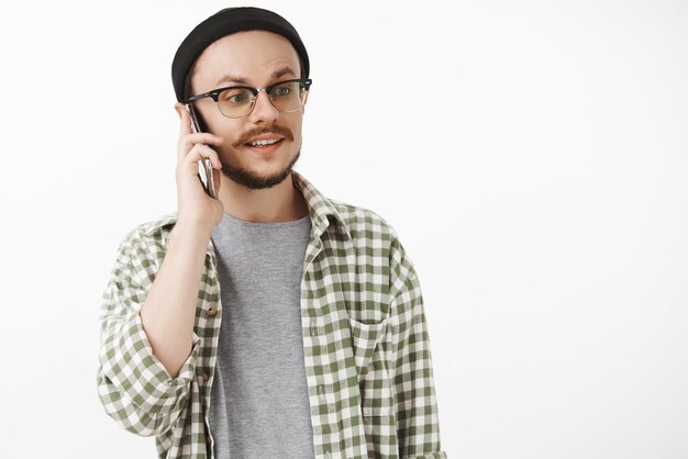 беззаботный, спокойный и расслабленный нормальный парень в шапочке и очках, который смотрит прямо с небрежным выражением лица во время разговора по телефону
