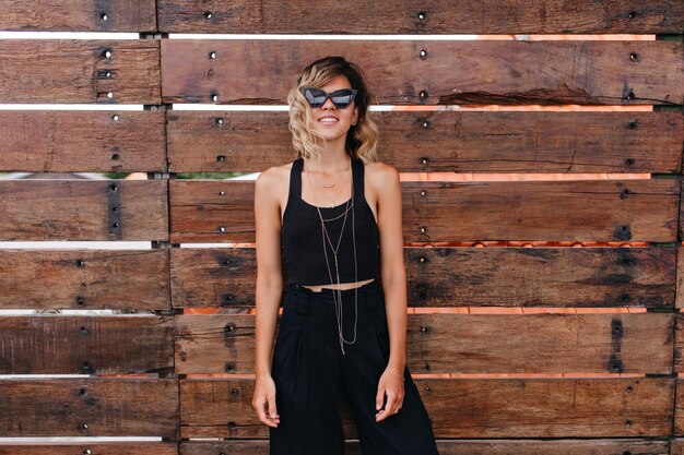 Carefree caucasian female model in black clothes standing near wooden wall. relaxed young woman with blonde hair enjoying photoshoot.