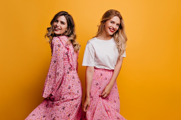 Carefree caucasian woman in pink skirt posing  Fascinating sisters standing on yellow wall with smile.