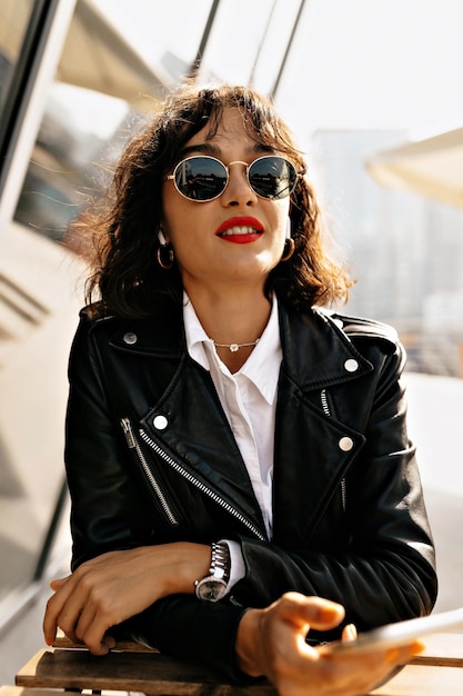 Carefree caucasian woman with red lips enjoying city views in warm windy day Outdoor photo of cute female lady in trendy jakcet drinking coffee on summer terrace in european town