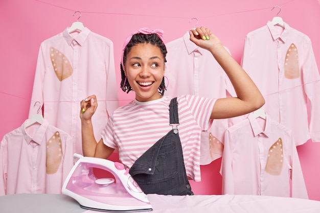 Carefree cheerful Afro American woman with dreadlocks and dark skin dances near ironing board keeps arms up smiles broadly happy to finish housework in time poses against shirts on hangers around