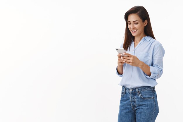 Carefree cheerful female entrepreneur waiting take-away coffee, order lunch online app, smiling delighted, hold smartphone, read text message phone screen, stand white wall upbeat