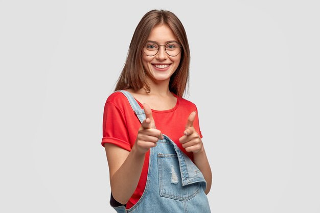 Carefree cheerful young woman with dark long hair, makes gun gesture with both index fingers, has charming smile