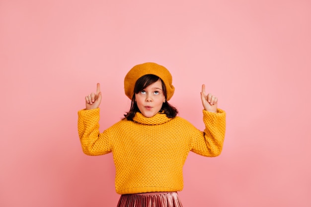 Carefree child in yellow beret looking up. Caucasian kid making funny faces on pink wall.