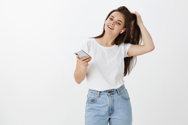 Carefree confident young woman posing with her phone and earbuds against white wall