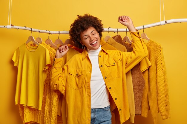 のんきな巻き毛の女性の服のバイヤーは幸せで踊り、腕を上げ、新しいコレクションから黄色の服を購入し、成功したショッピングの日を喜び、元気で、ラックの衣装と踊ります