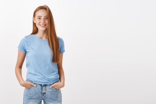 Foto gratuita spensierato carino e timido felice giovane ragazza rossa con le lentiggini sorridendo gioiosamente tenendosi per mano in tasca in piedi in posa ordinaria casual amichevole e gioioso