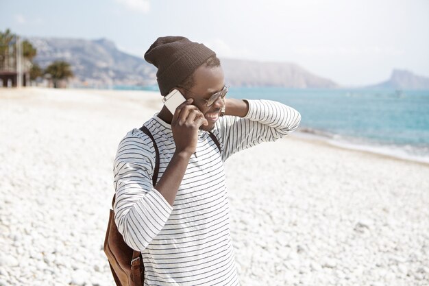 Carefree dark-skinned hipster in trendy clothes having conversation on smartphone while walking along pebble beach, relaxing on summer day by sea
