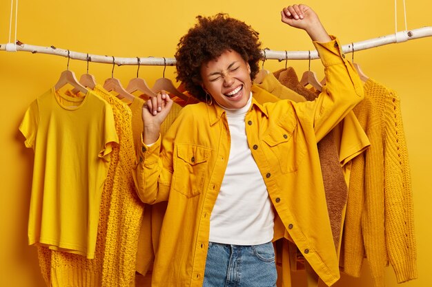 Carefree dark skinned woman dances in rhythm of music, makes victory movement, dressed in yellow shirt and jeans, moves against rack filled of fashionable clothes