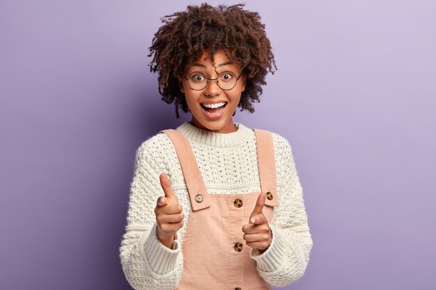 Carefree dark skinned young woman points directly, makes finger gun gesture, looks happily, expresses good emotions, wears casual clothes, isolated against purple wall. Bang bang