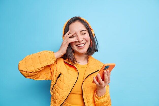 Carefree fashionable Asian woman smiles positively keeps eyes closed hand on face has fun listens music holds modern smartphone wears wireless headphones expresses positive emotions poses indoor