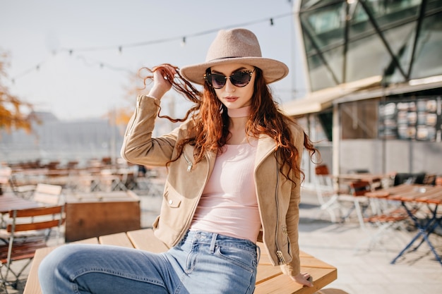 Carefree female model in dark glasses posing in hat in street cafe