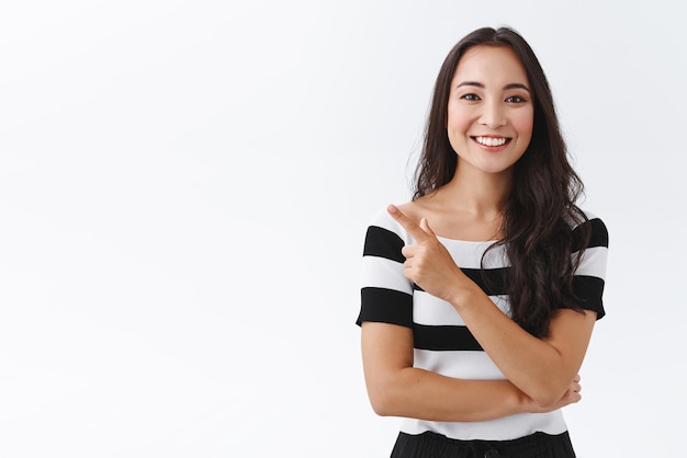 Carefree friendlylooking young woman in striped tshirt with joyful entertained gaze pointing upper left corner and smiling broadly curious about interesting promo stand white background