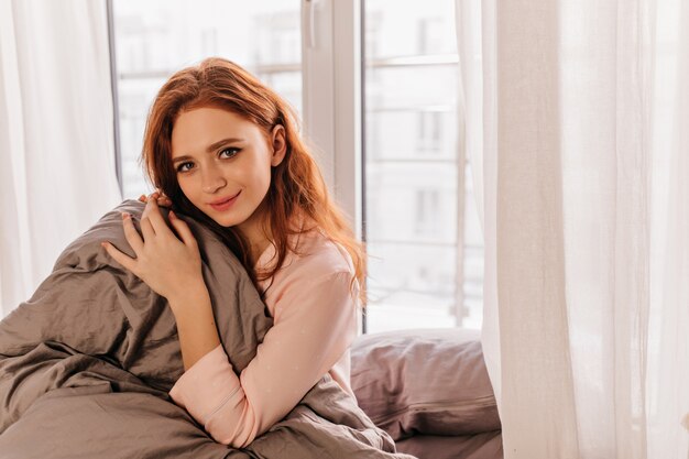 ベッドに座っているかわいいパジャマでのんきな女の子。朝に冷える素敵な生姜の女性。