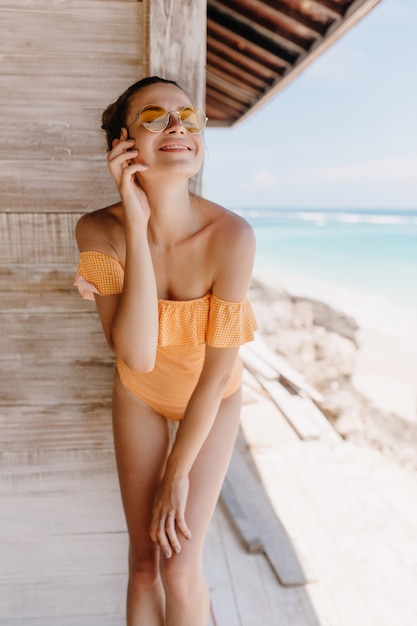 Беззаботная девушка позирует на пляже с закрытыми глазами и искренней улыбкой. Открытый снимок великолепной стройной дамы в оранжевом купальнике.
