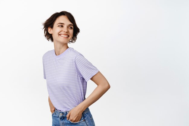 Carefree girl, posing on isolated white, turn behind shoulder, laughing and smiling happy, hands in pockets, looking at empty copy space