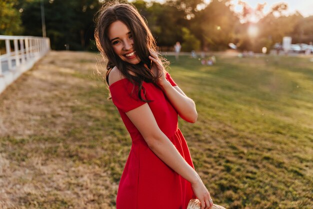 Carefree girl in stylish red dress dancing in sunny day. Enthusiastic brunette young woman in summer attire chilling in park.