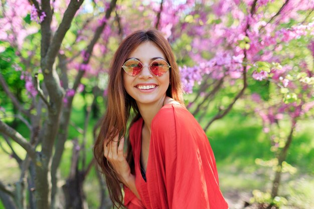 Carefree girl in stylish straw hat and coral dress enjoying spring day in sunny garden on blooming tree