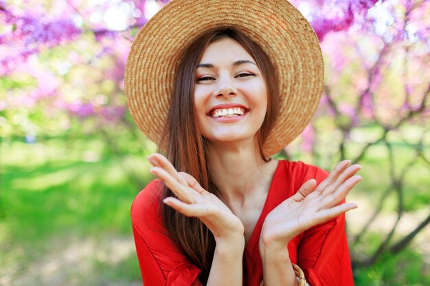 Carefree girl in stylish straw hat and coral dress enjoying wale spring day in sunny garden on blooming tree