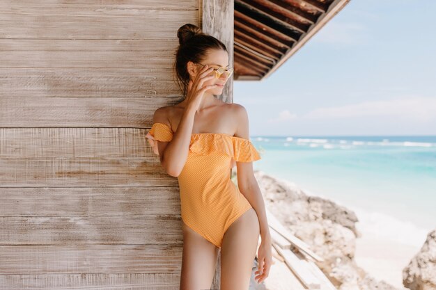 Carefree girl in vintage swimwear standing near wooden house and looking at sea. Outdoor photo of gorgeous brunette woman with trendy hairstyle relaxing at resort.