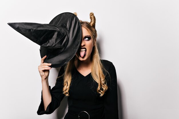 Carefree girl in witch costume making faces on white wall. Positive female vampire holding black hat during halloween photoshoot.