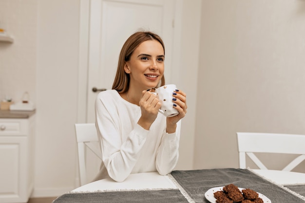 キッチンでコーヒーを飲みながらのんきな女の子。コーヒーブレイクを楽しんでいる白いシャツを着た楽しい笑顔の女性の写真。