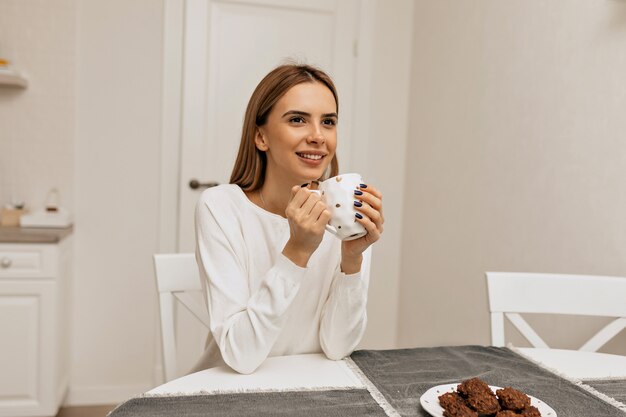 Carefree girl with drinking coffee at kitchen. Photo of pleasant smiling woman in white shirt enjoying coffee break.