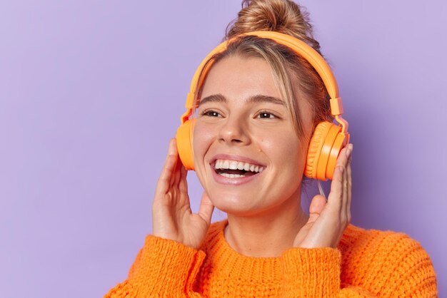 Carefree glad young woman smiles broadly keeps hands on stereo headphones with good sound quality enjoys favorite song dressed in knitted jumper isolated over purple background. Leisure concept